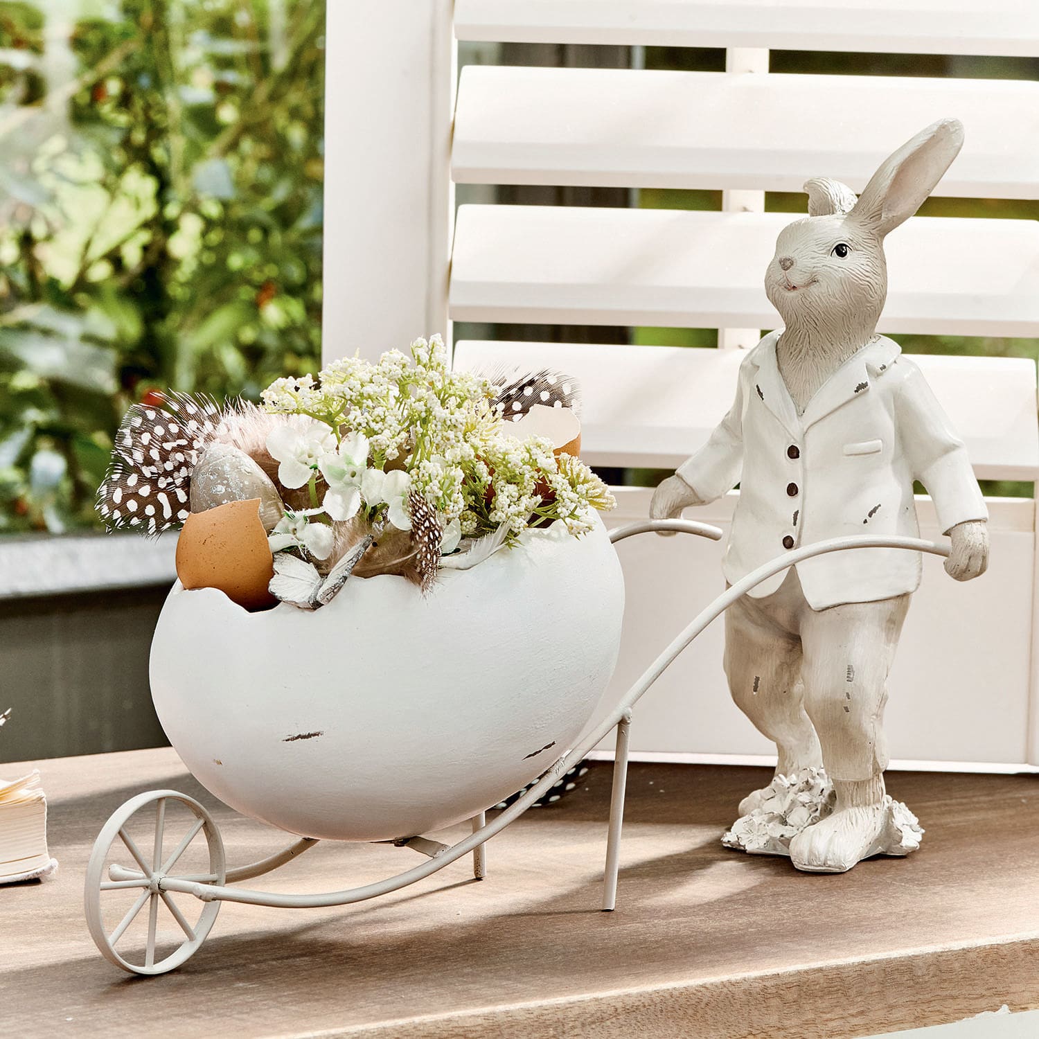 Hase Jaris Dekorativer weisser Hase mit Keramikei und Blumen in Metallwagen auf dem Tisch.