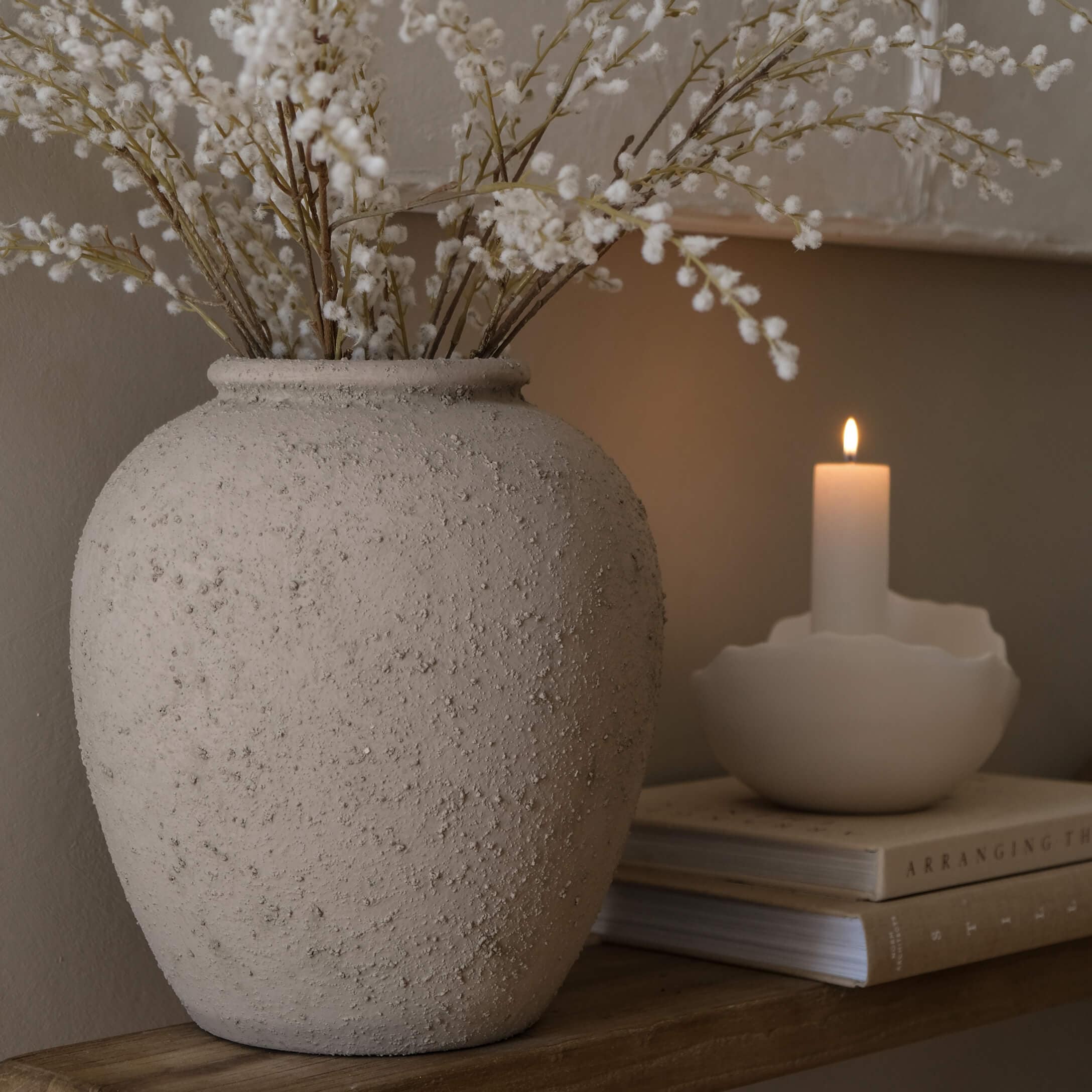 Beige strukturierte Keramikvase mit getrockneten weißen Blüten auf Holzregal neben brennender Kerze