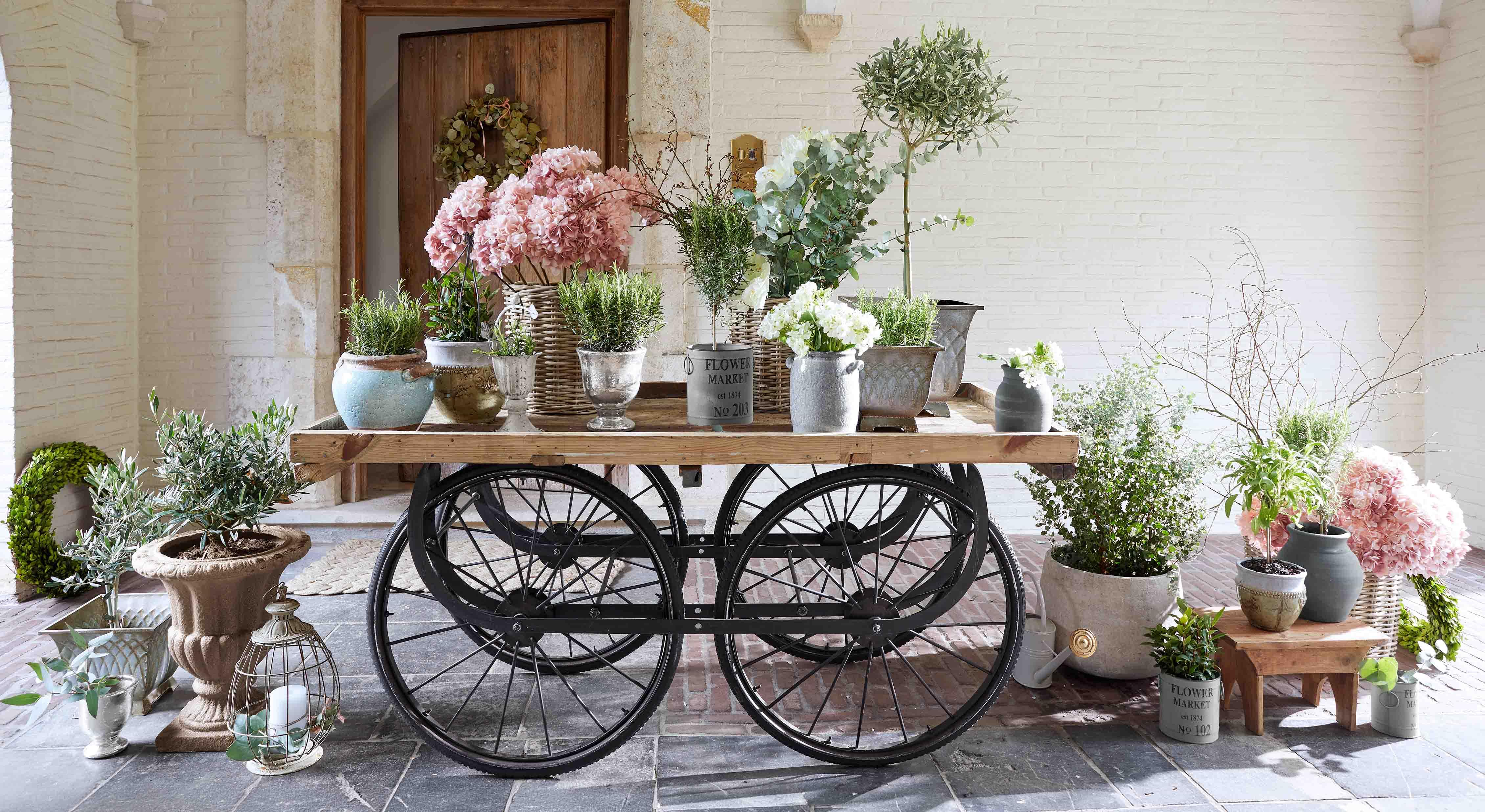 Holzwagen mit schwarzen Rädern und verschiedenen Blumentöpfen mit rosa und weißen Blumen
