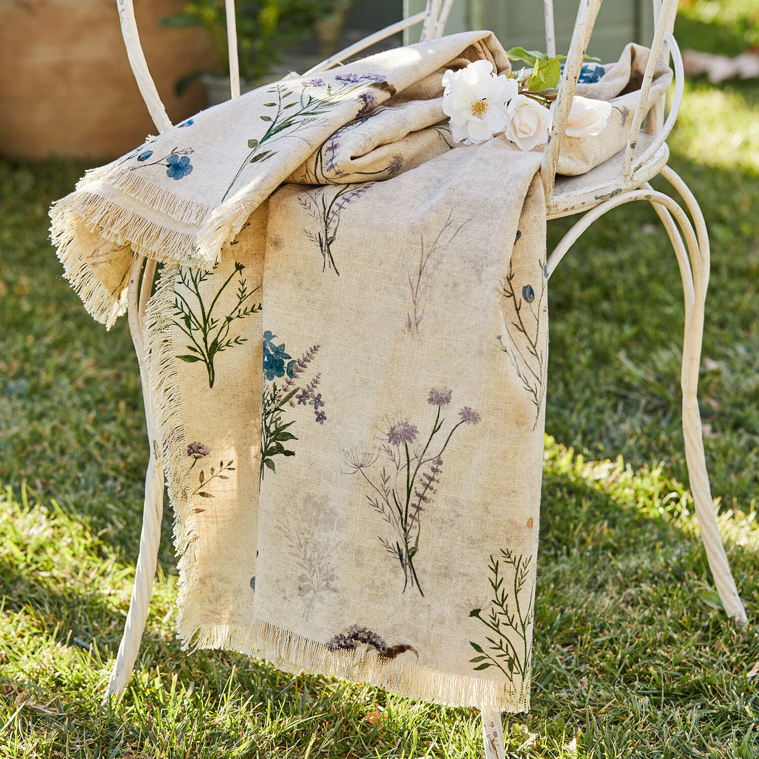 Plaid Cabinda Beige Leinentischdecke mit Blumenmuster und Fransen auf weissem Metallstuhl im Garten