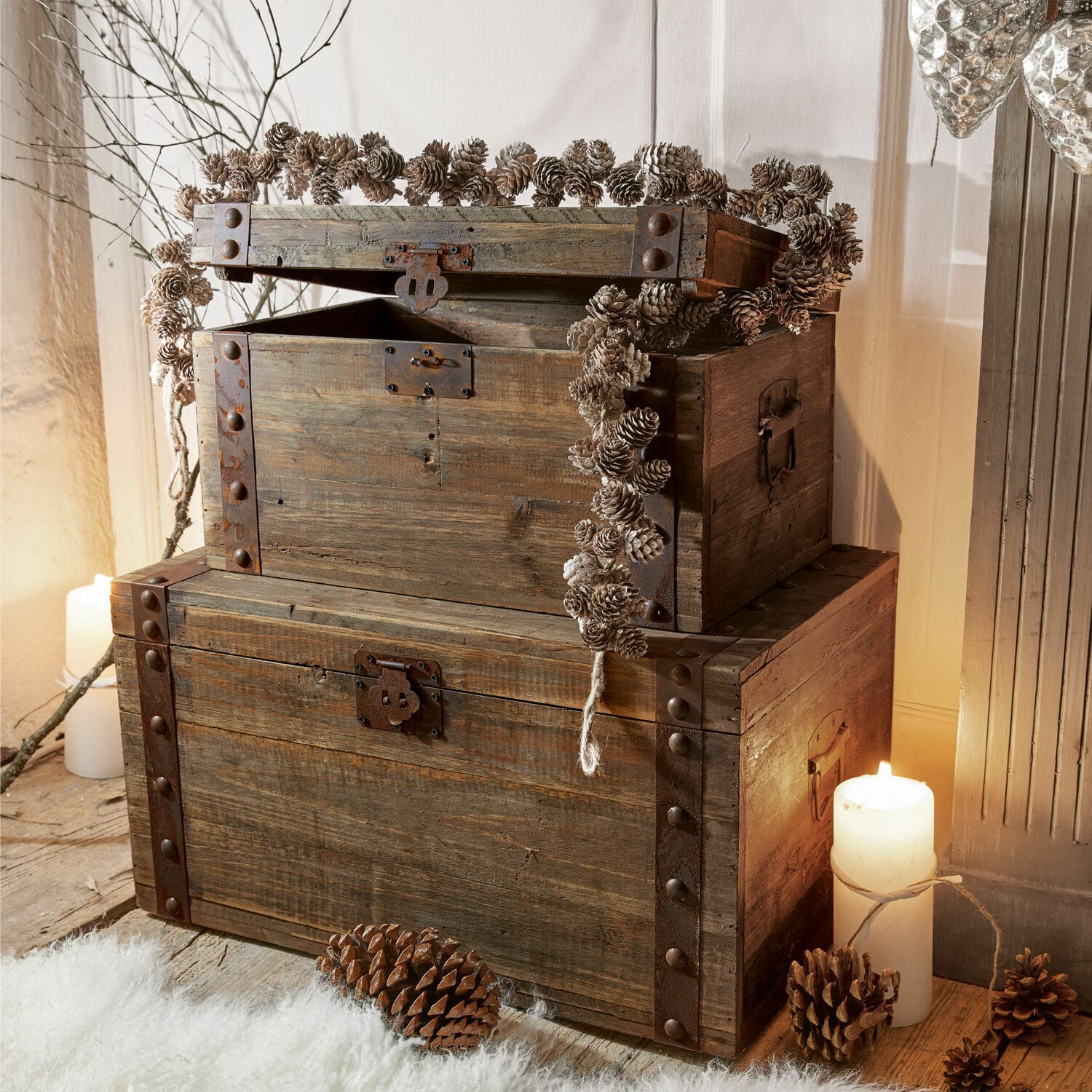 Truhe 2er Set Pratt Rustikale Holzkisten mit Metallbeschlägen, dekoriert mit Tannenzapfen und brennenden Kerzen.