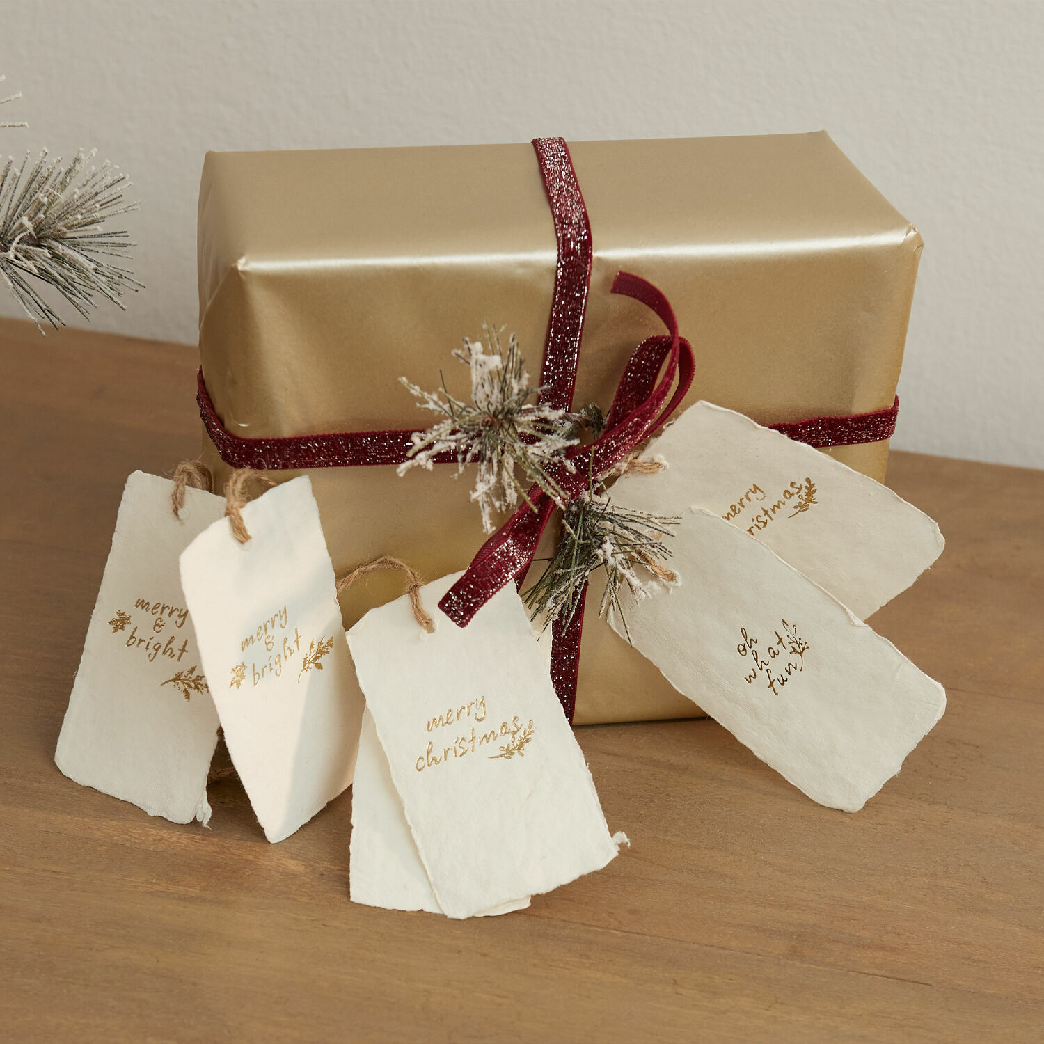 Anhänger 6er Set Aamira Goldverpacktes Geschenk mit bordeauxrotem Band und weissen Papieranhängern auf Holztisch