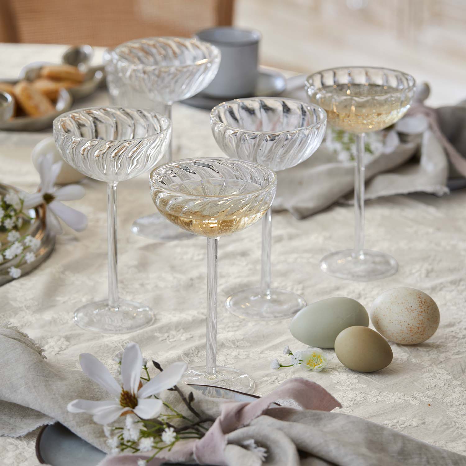 Fünf transparente Champagnergläser mit dekorativem Muster auf Tisch mit Stoff und Blumen