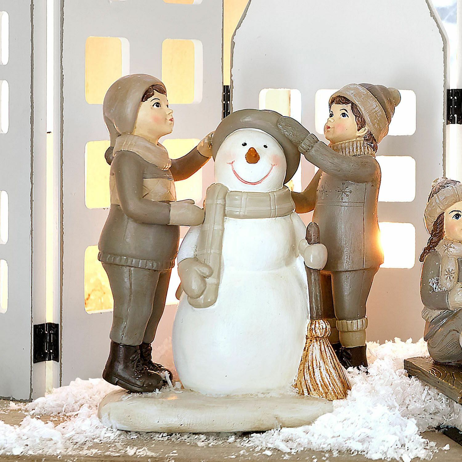 Deko-Figur Elvirette Figuren von Kindern, die einen Schneemann aus Material in Beige- und Brauntönen auf Dekoschnee anziehen.