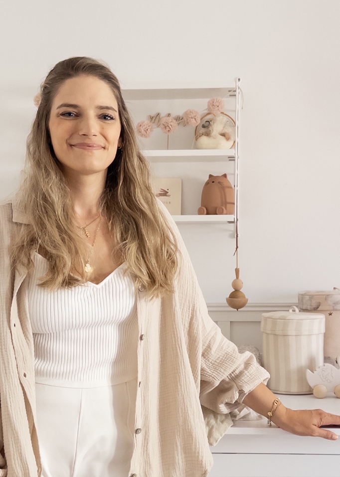 Lächelnde Frau in weissem Top und beiger Bluse in hellem, gemütlichem Raum mit Kinderdetails