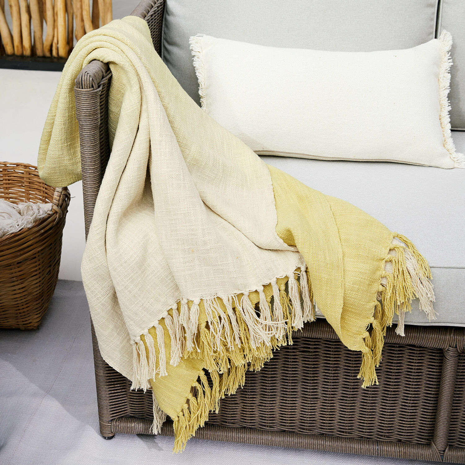 Plaid Aurice Zweifarbige Baumwoll-Decke in Creme und Gelb mit Fransen auf grauem Rattansofa
