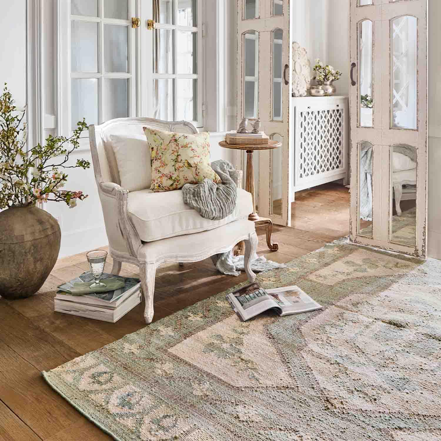 Weißer Holzsessel mit floralem Kissen in hellem Wohnzimmer mit Teppich und Spiegel im Vintage-Stil