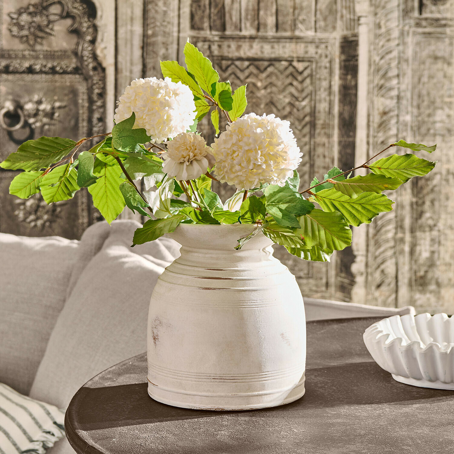 Vase Niverana Weisse Keramikvase mit grünen Blättern und cremefarbenen Blüten auf dunklem Vintage-Tisch